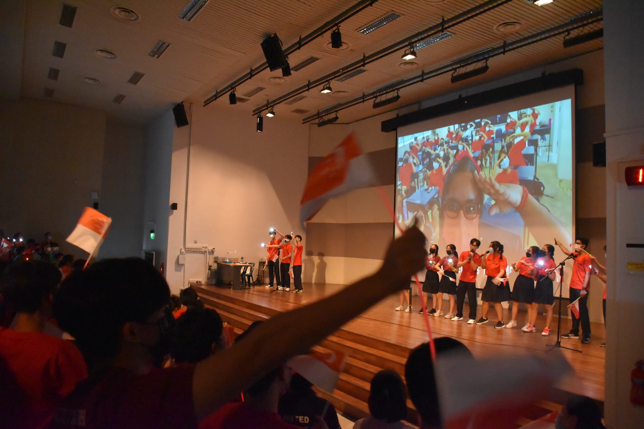 Students in red shirts perform on a stage while the audience waves red and white flags in an indoor hall.